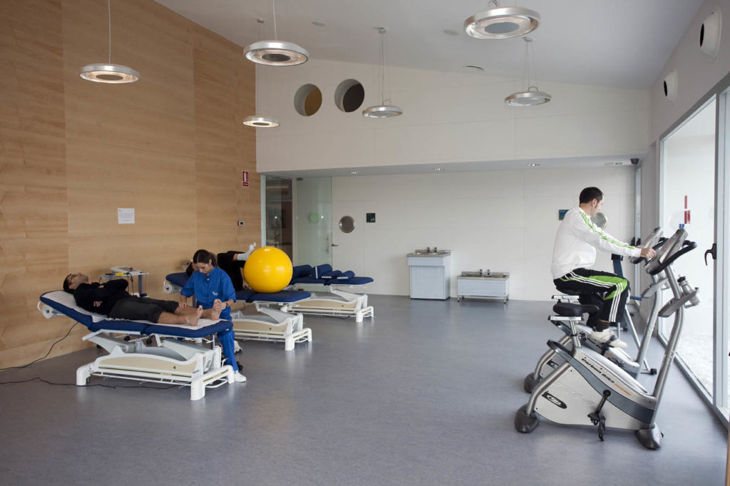 Foto de dos personas en el gimnasio de rehabilitación con una persona siendo atendida y otra utilizando una bicicleta estática